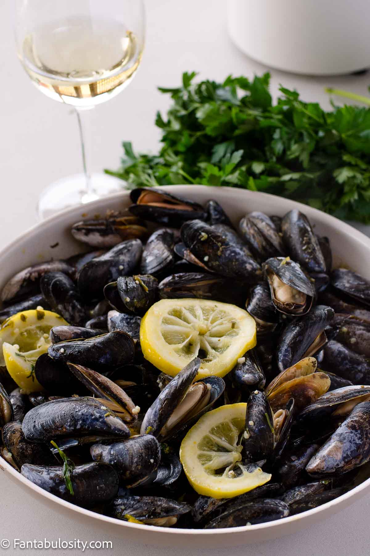 Cooked mussels in pan with white wine sauce, and lemon slices on top, next to a glass of wine.