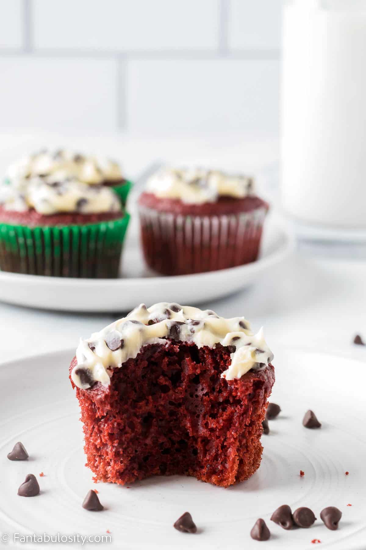 Red velvet cupcake with chocolate chip frosting, with a bite taken out of it, sitting on a white plate.