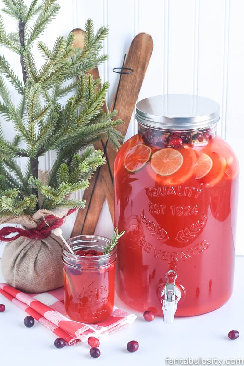 Fruit Punch in glass with rosemary sprig and large batch in a drink dispenser.