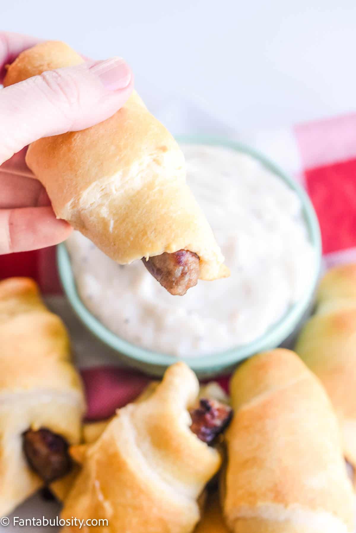 hand holding breakfast pigs in a blanket over bowl of gravy