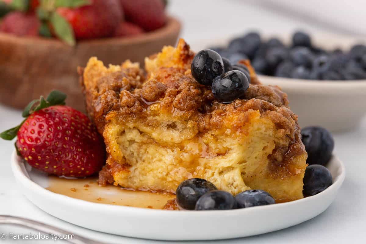 Slice of french toast casserole on white plate with syrup and berries.