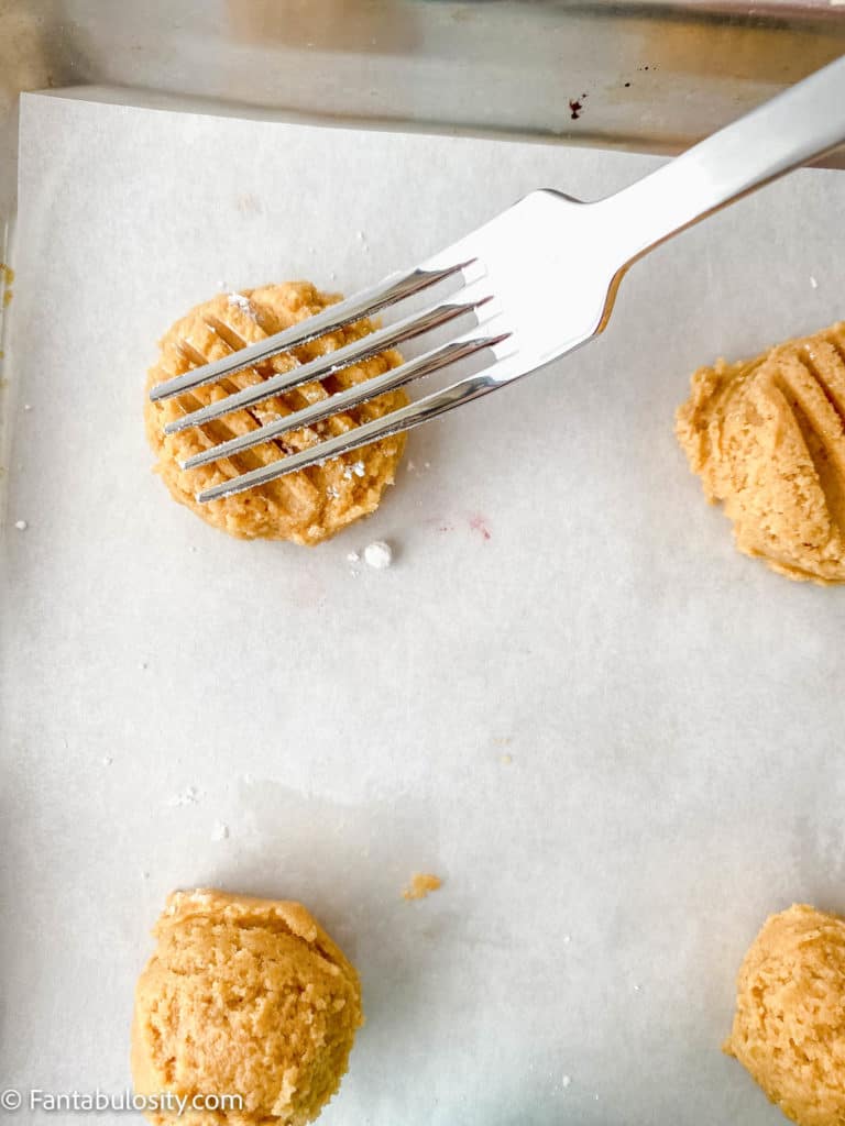 A fork flattening a peanut butter cookie dough ball to create a criss-cross pattern.