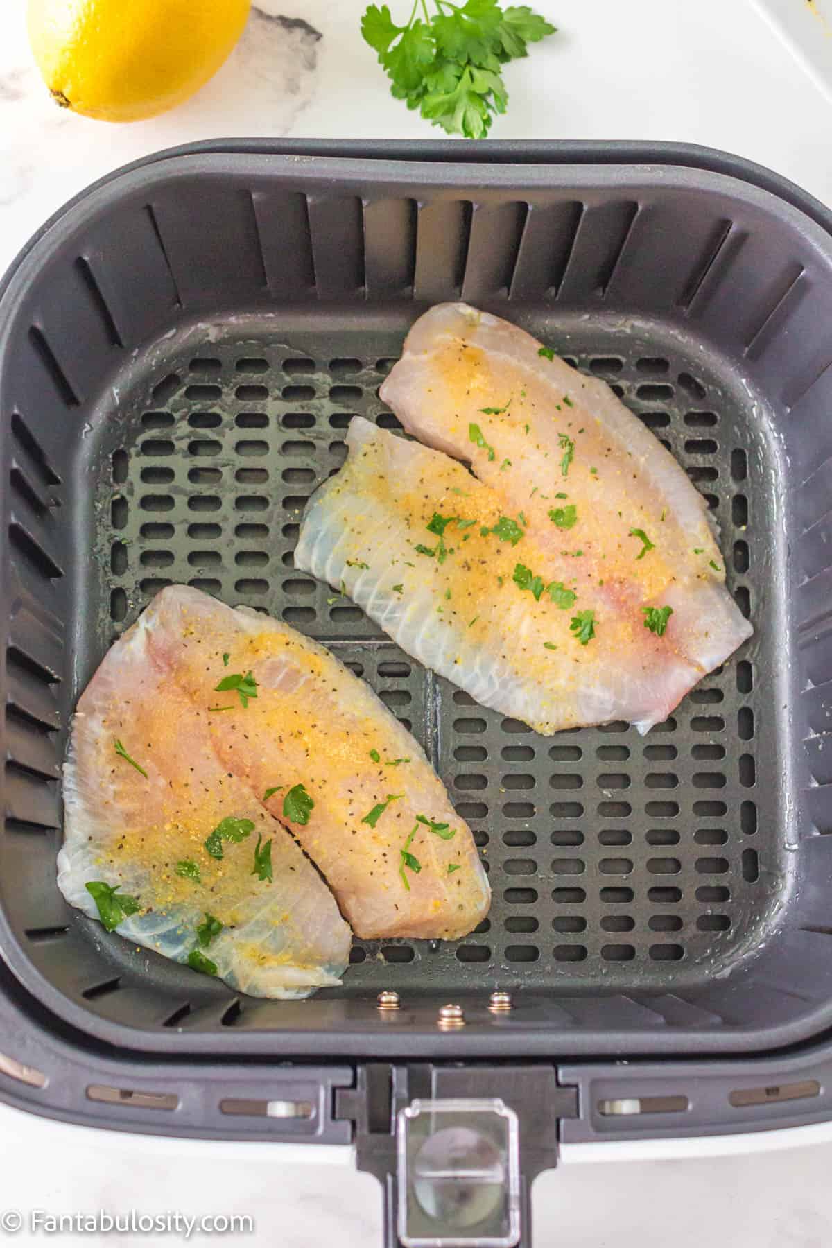 Raw tilapia in then air fryer basket, covered in spices.