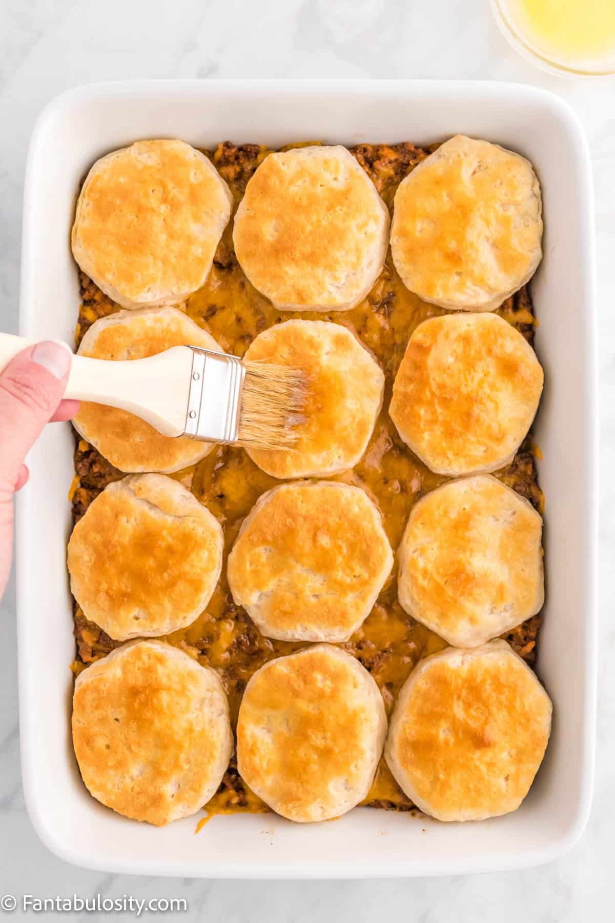 A pastry brush brushing melted butter on top of the biscuits after the cheeseburger casserole has been cooked.