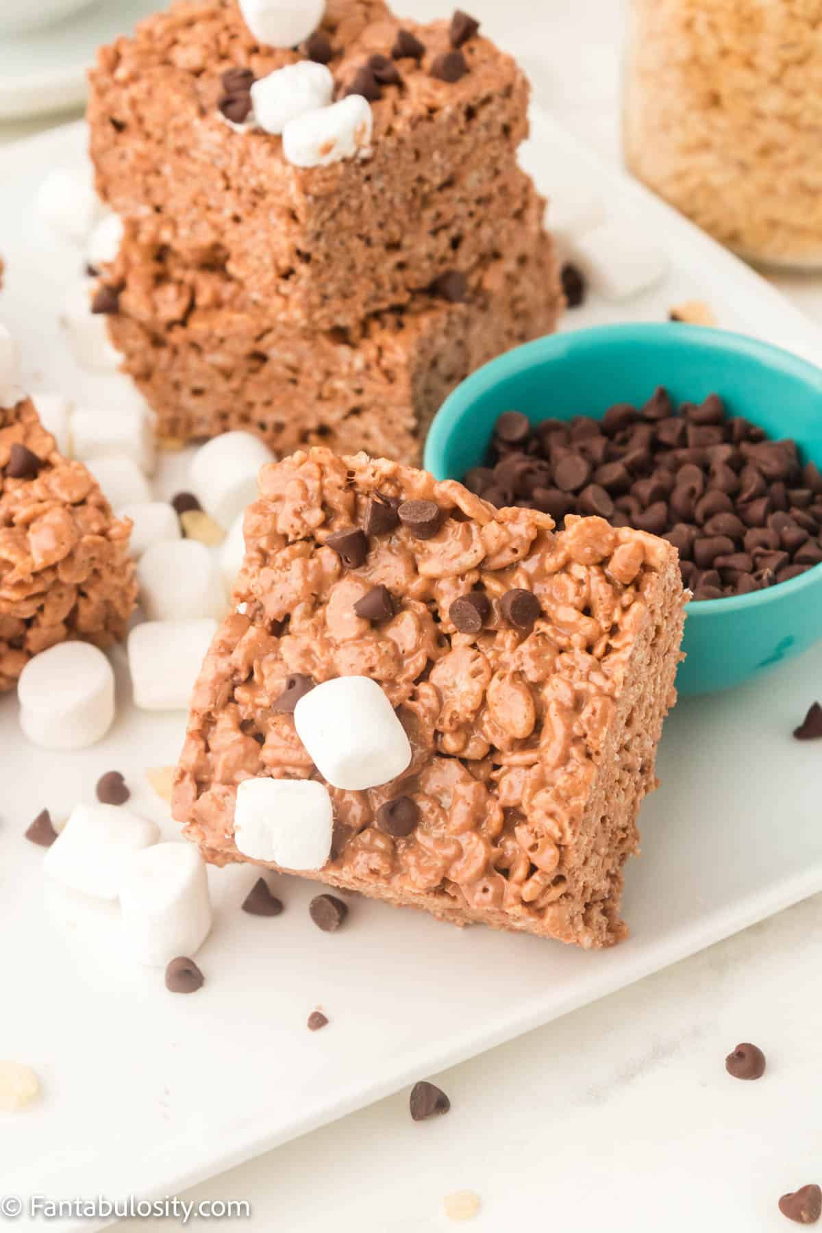 Chocolate Rice Krispie treats on a tray with a bowl of chocolate chips beside them.