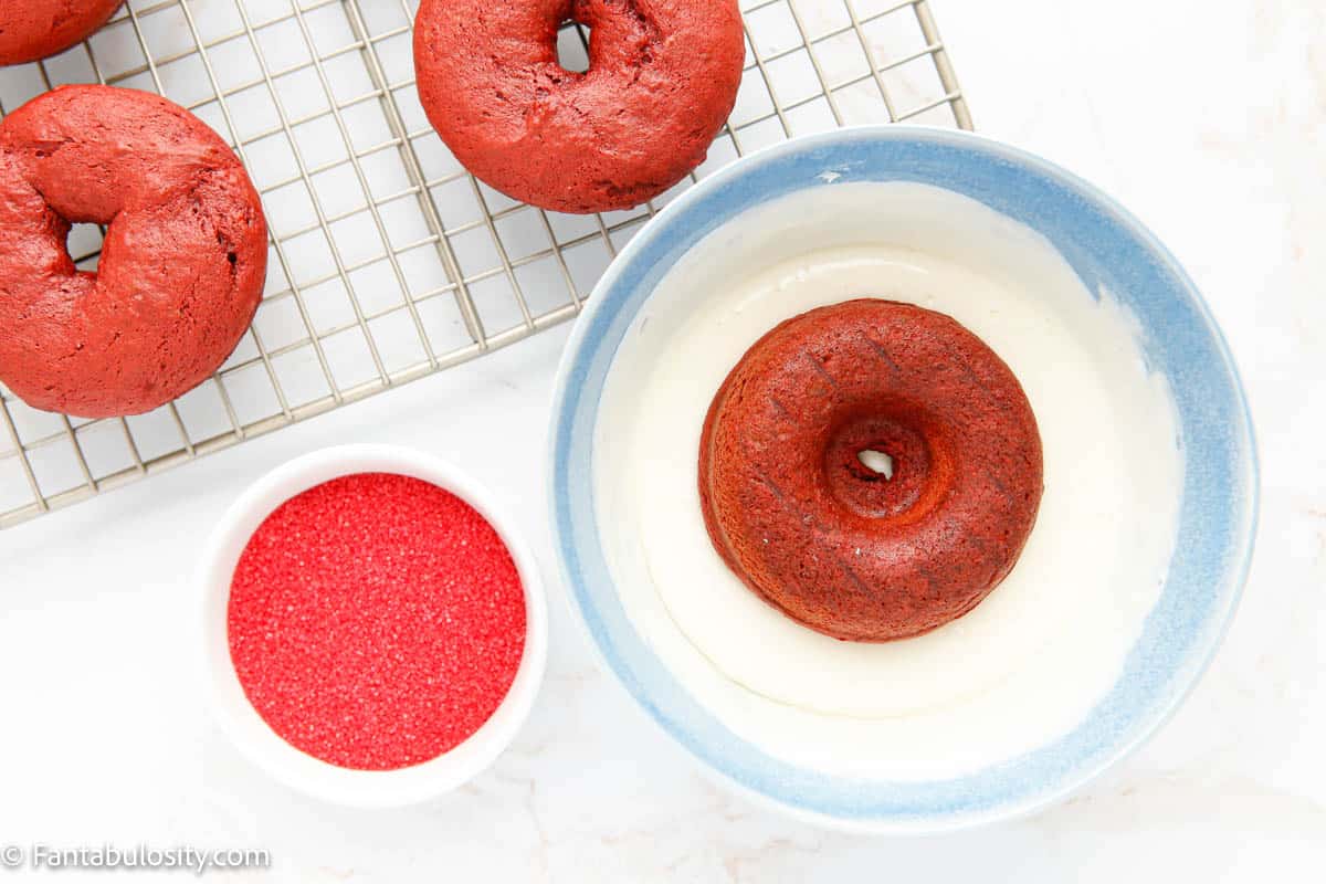 A red velvet donut being dipped in a bowl of cream cheese icing with a bowl of red sprinkles beside it.