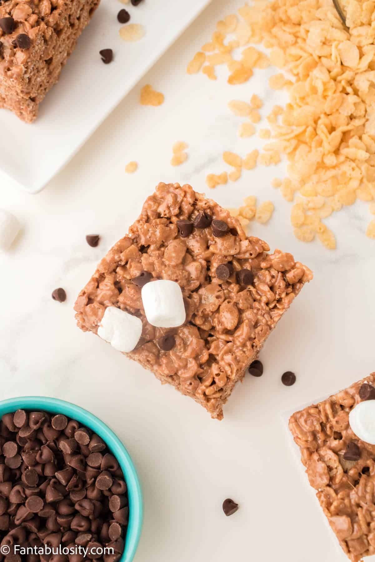Chocolate Rice Krispie Treats with chocolate chips and Rice Krispies on the table next to them. 