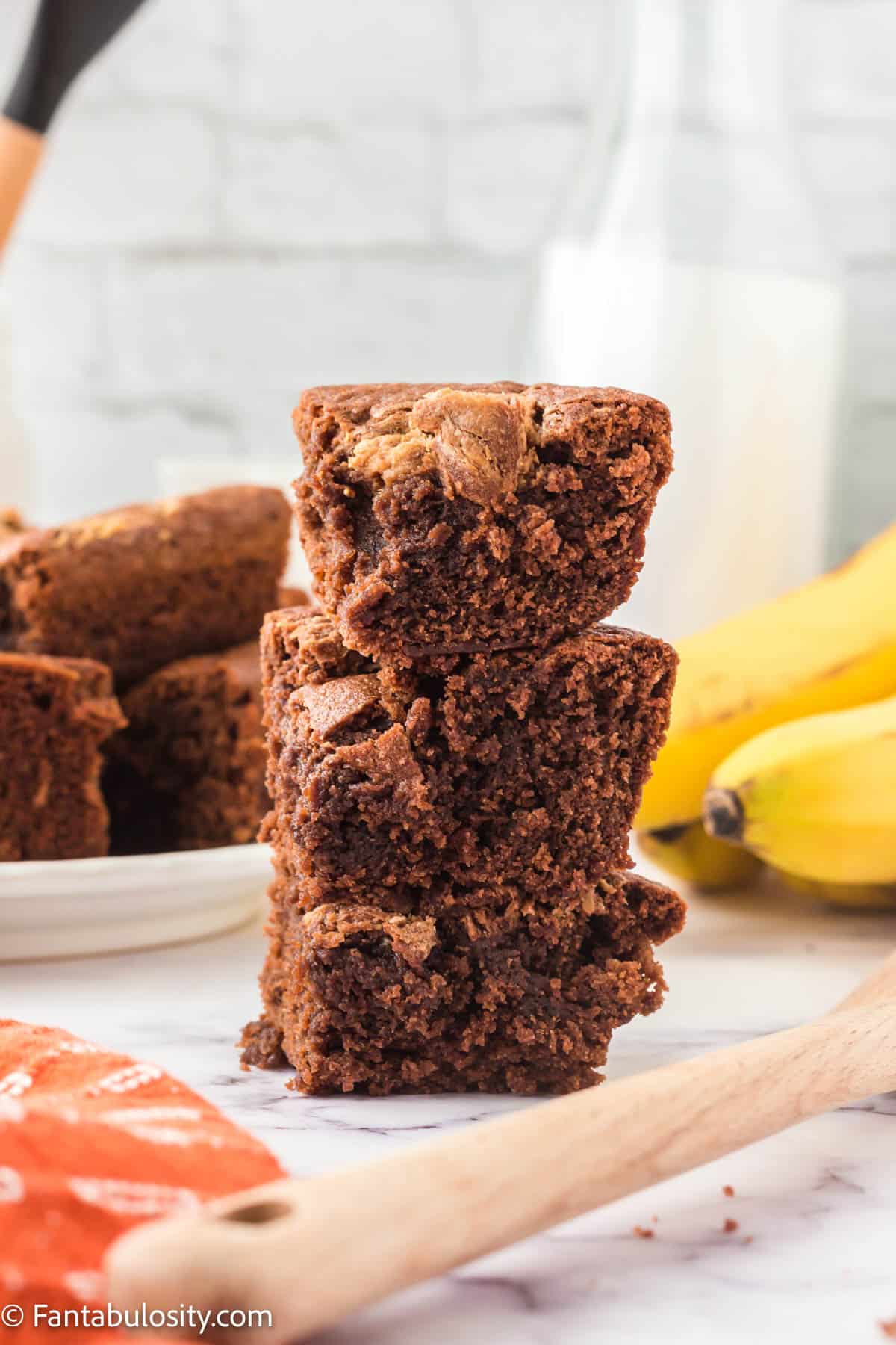 A stack of brownies with a plate of brownies and bunch of bananas in the background.