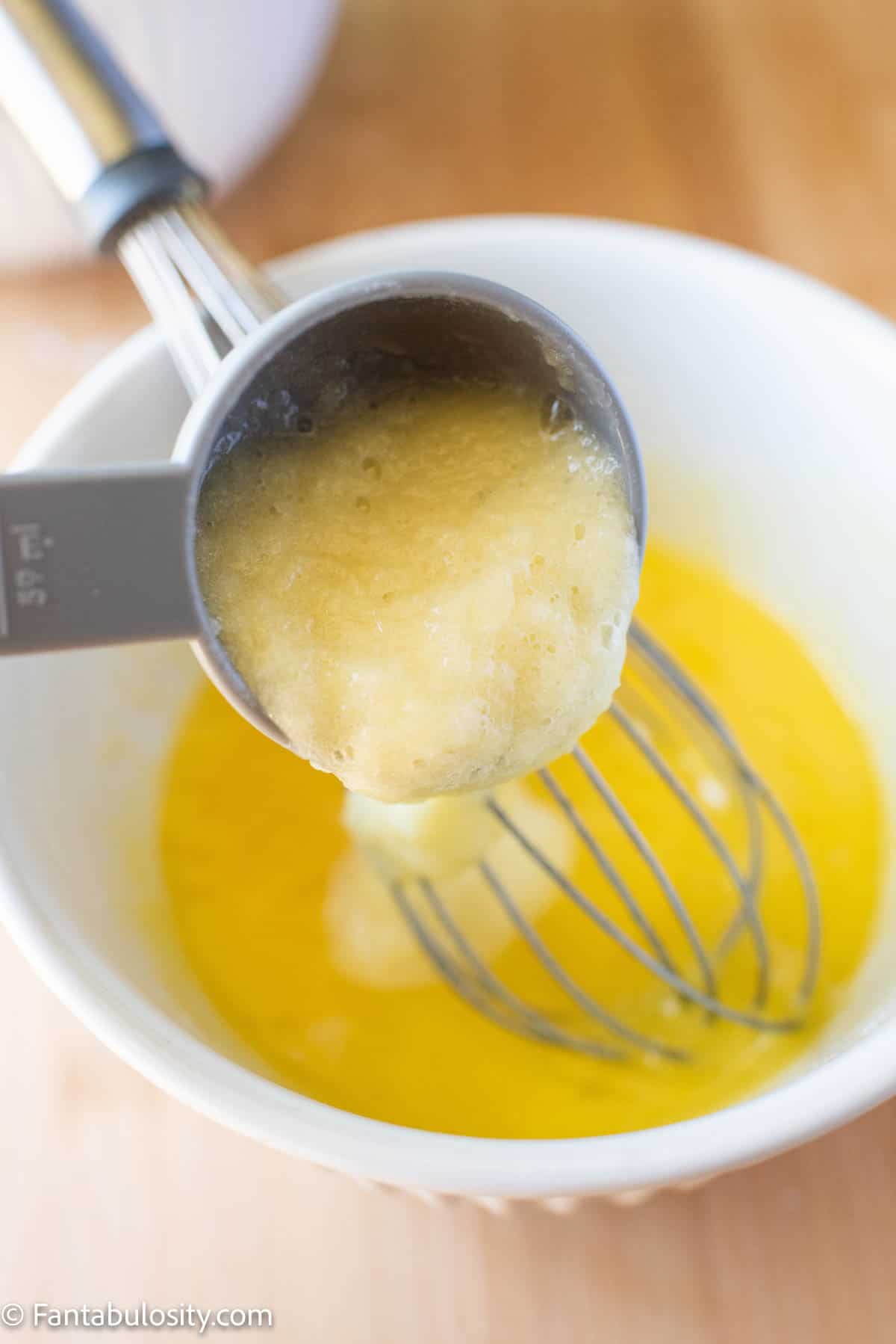 A measuring cup pouring butter into a bowl with liquid and a whisk in it.