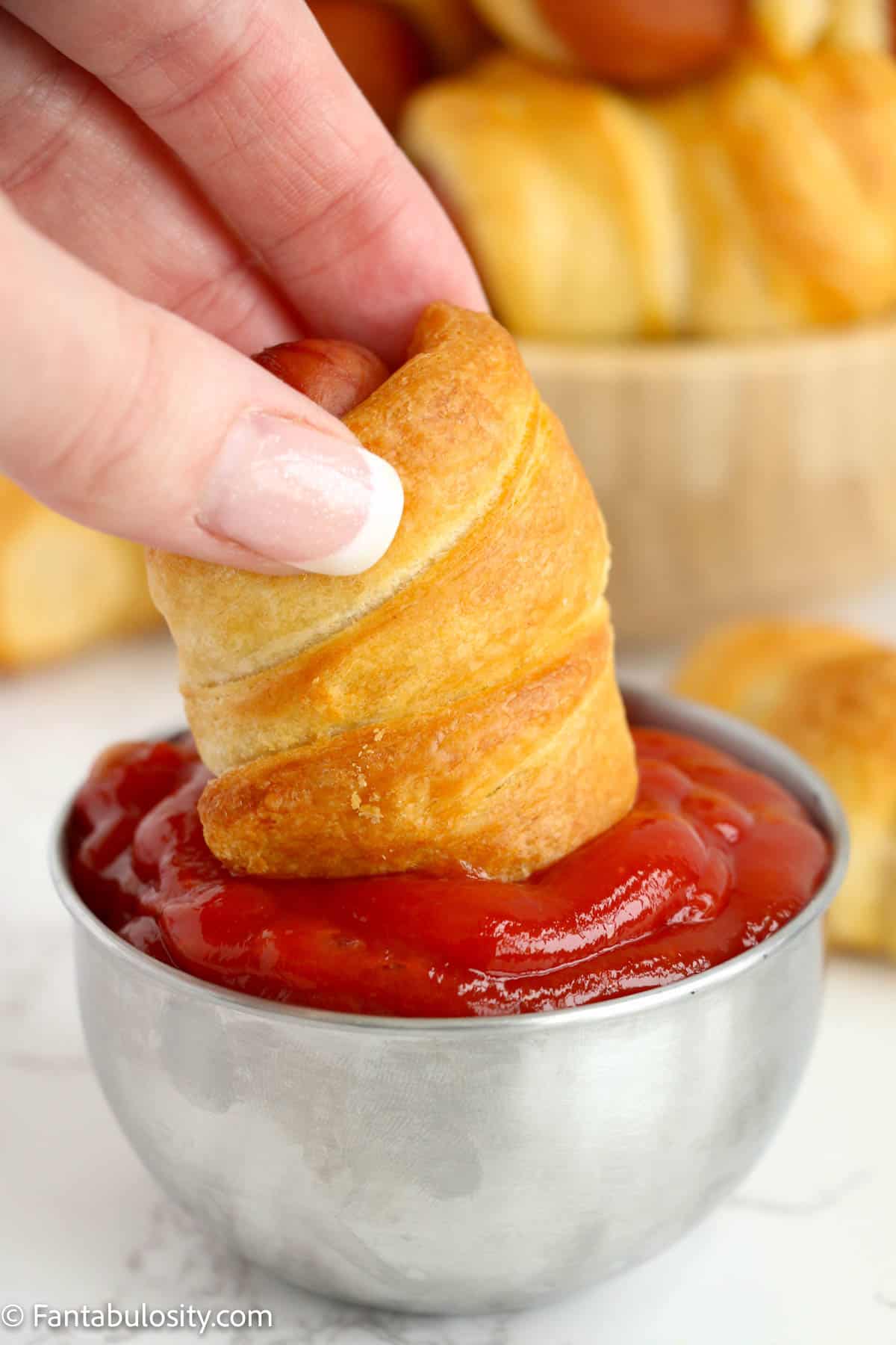 Someone dipping a pig in a blanket into ketchup that is in a tiny stainless steel bowl.