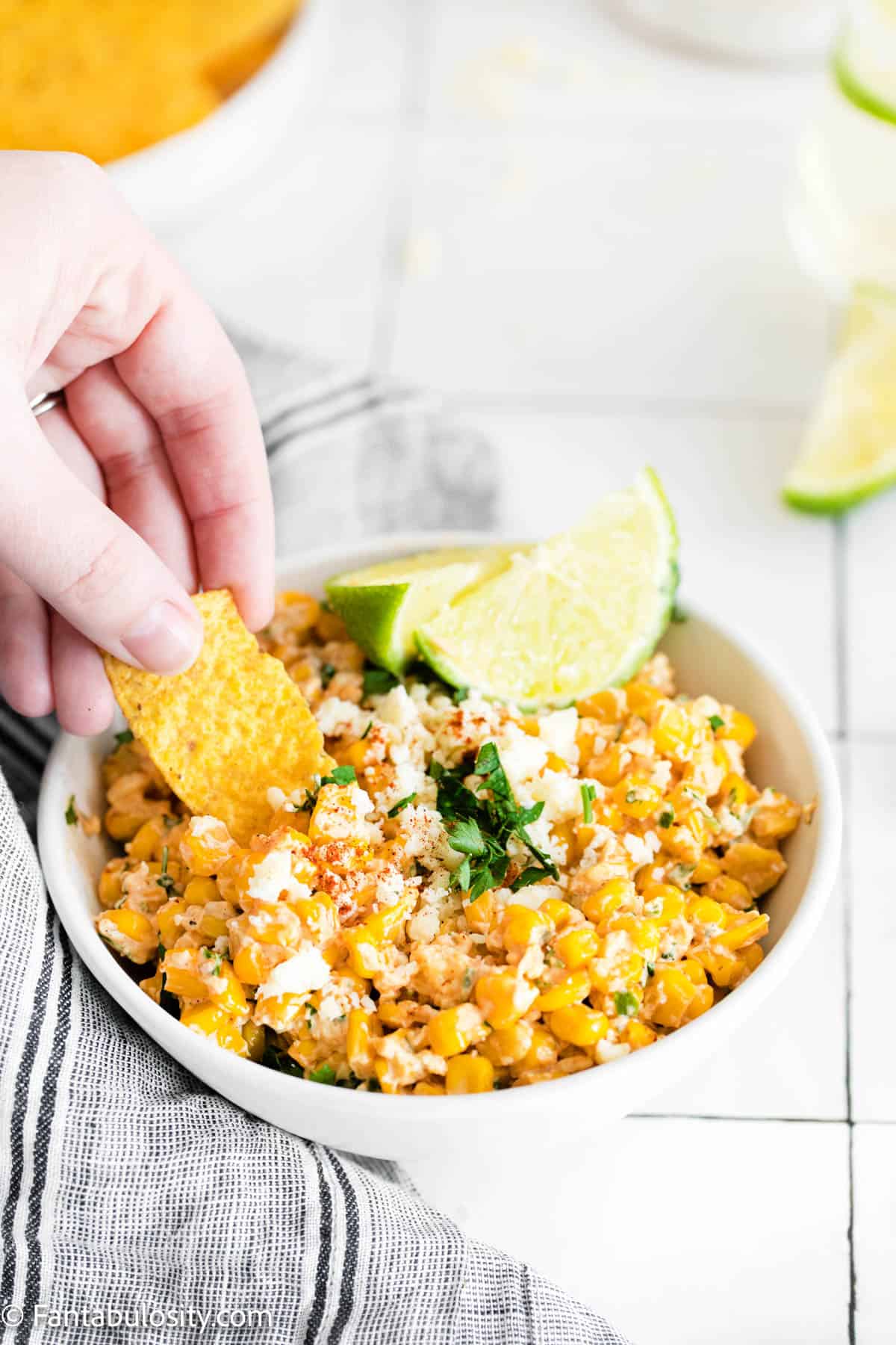Elote corn dip in a bowl with lime wedges and someone dipping a corn chip into it.