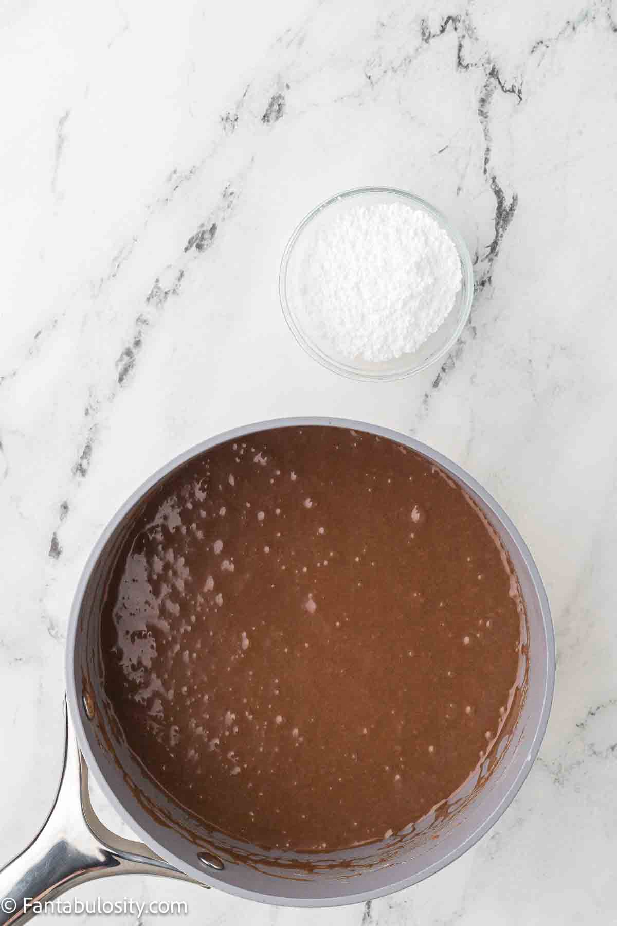 Powdered sugar sitting next to chocolate frosting mixture in sauce pan. 