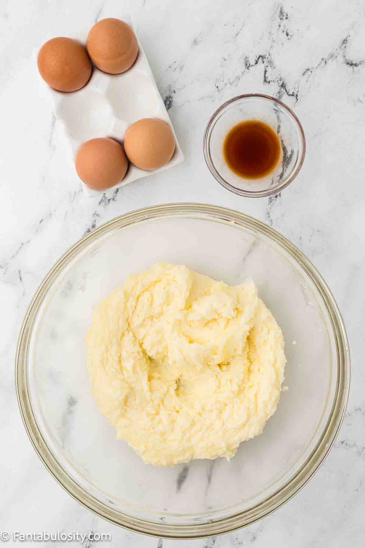 Butter and sugar creamed together in mixing bowl, with eggs and vanilla sitting on the side.