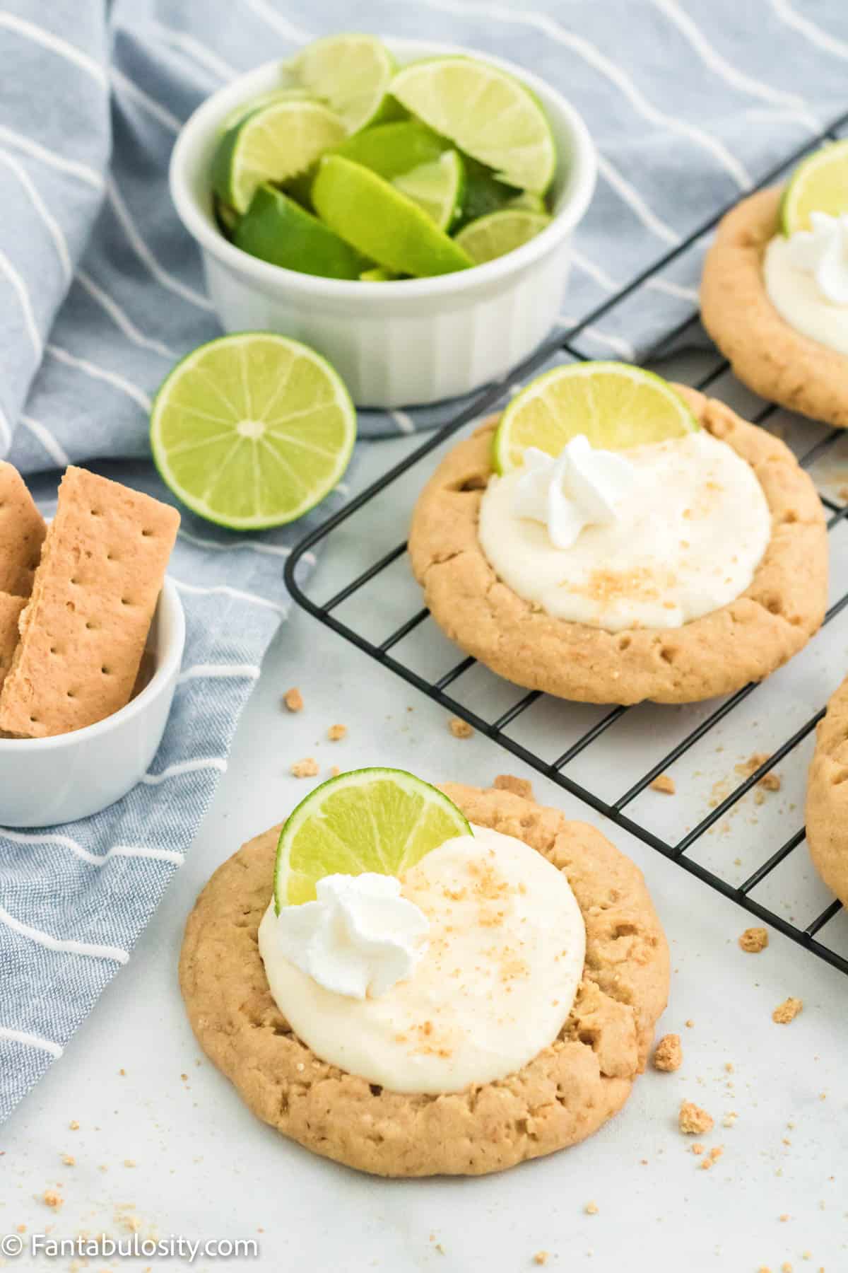 Key lime pie cookies on a wire rack with a bowl of graham crackers and a bowl of key limes next to them. 