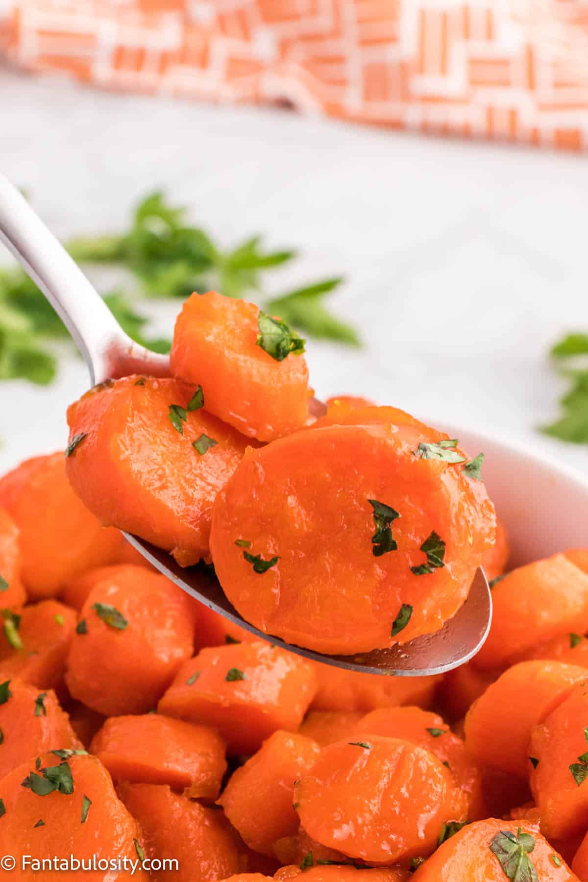 A spoon filled with cooked carrots is displayed in front of a bowl of cooked Candied Carrots