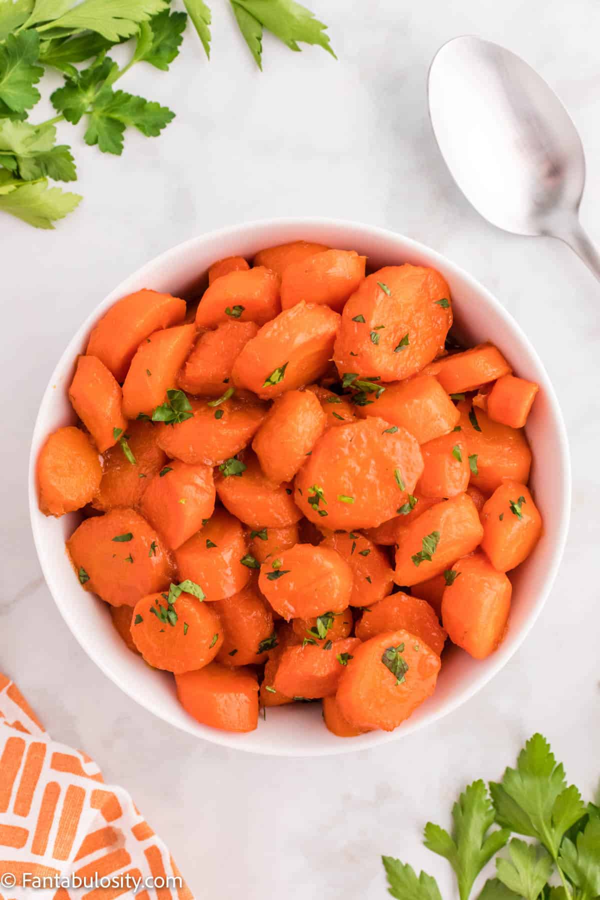 Cooked Candied Carrots are shown in a white serving bowl with a spoon in the upper right hand corner and an orange patterned napkin in the bottom left corner