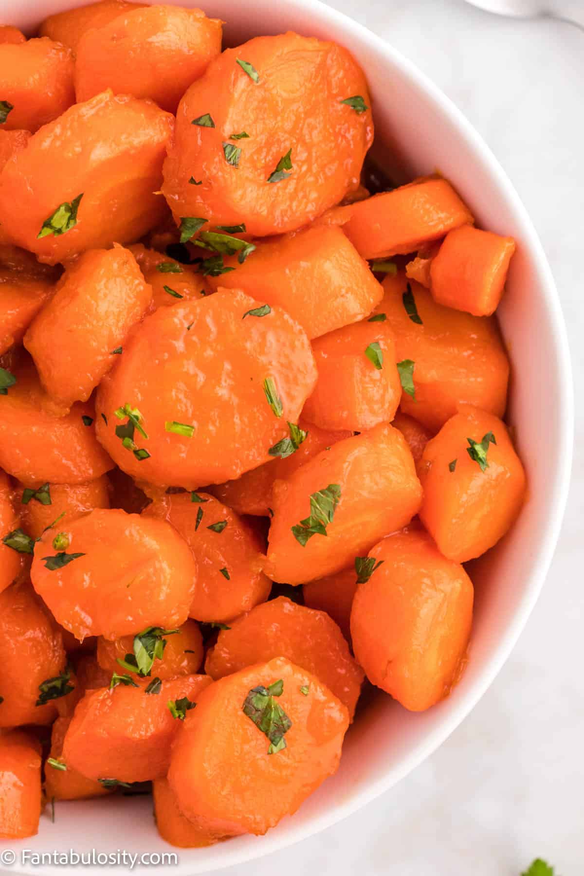 Close up up cooked candied carrots topped with bright green parsley in a white serving bowl
