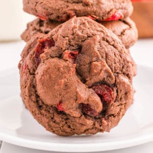 A chocolate cherry cookie on a plate leaning against a stack of cookies.