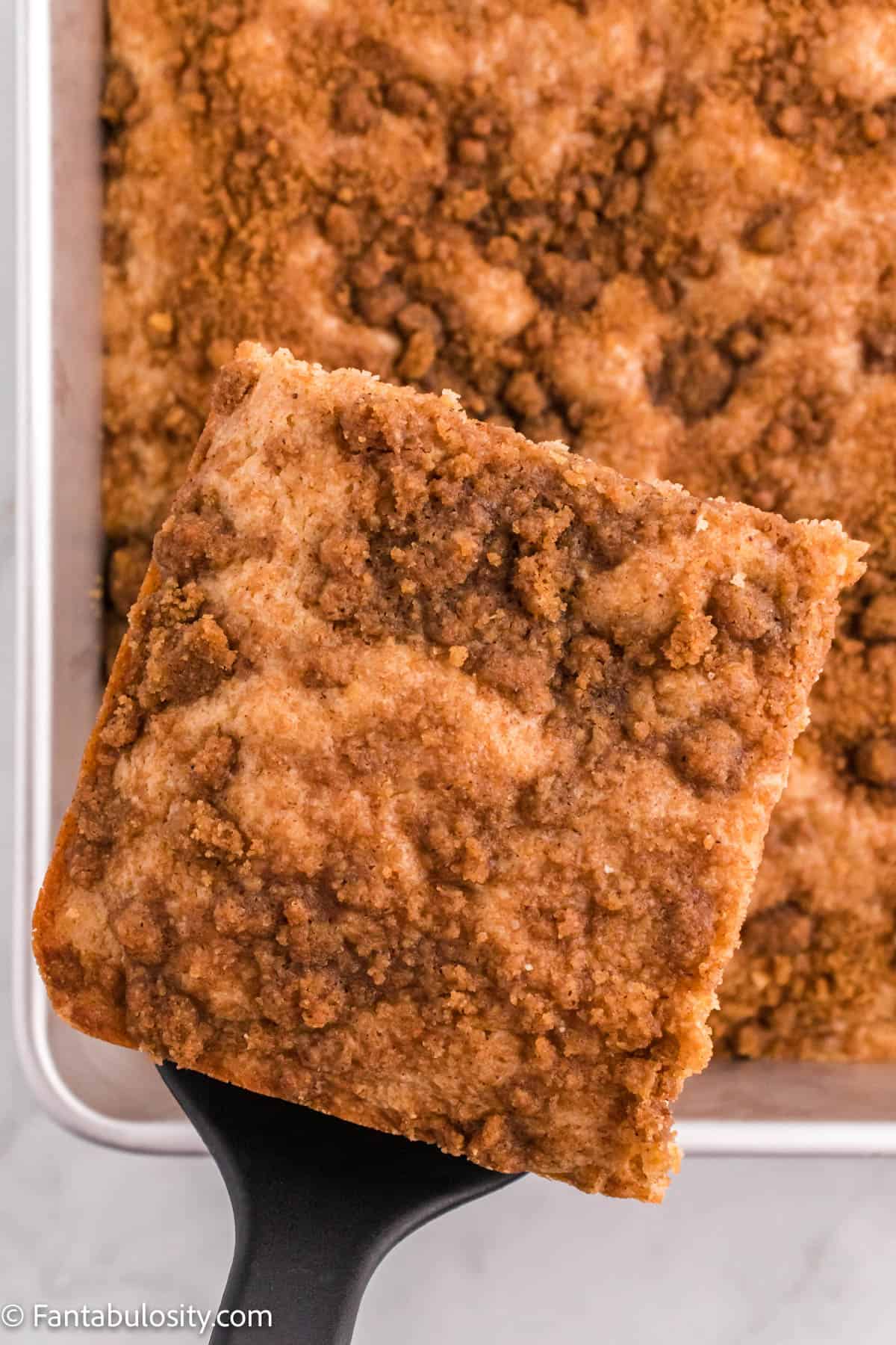 A slice of Cinnamon Coffee Cake is displayed on a spatula above the cake pan