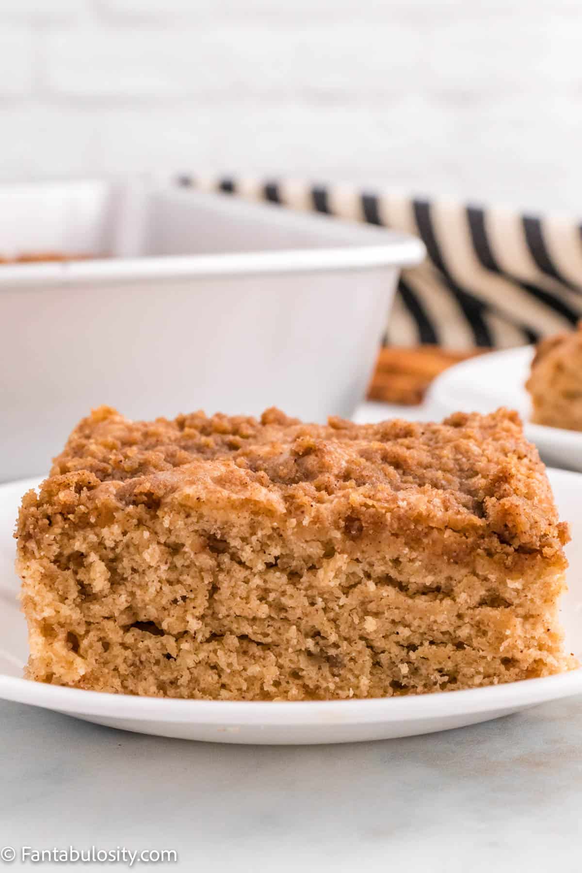 A slice of Cinnamon Coffee Cake on a white plate is in the center of the photo with the pan of cake in the background