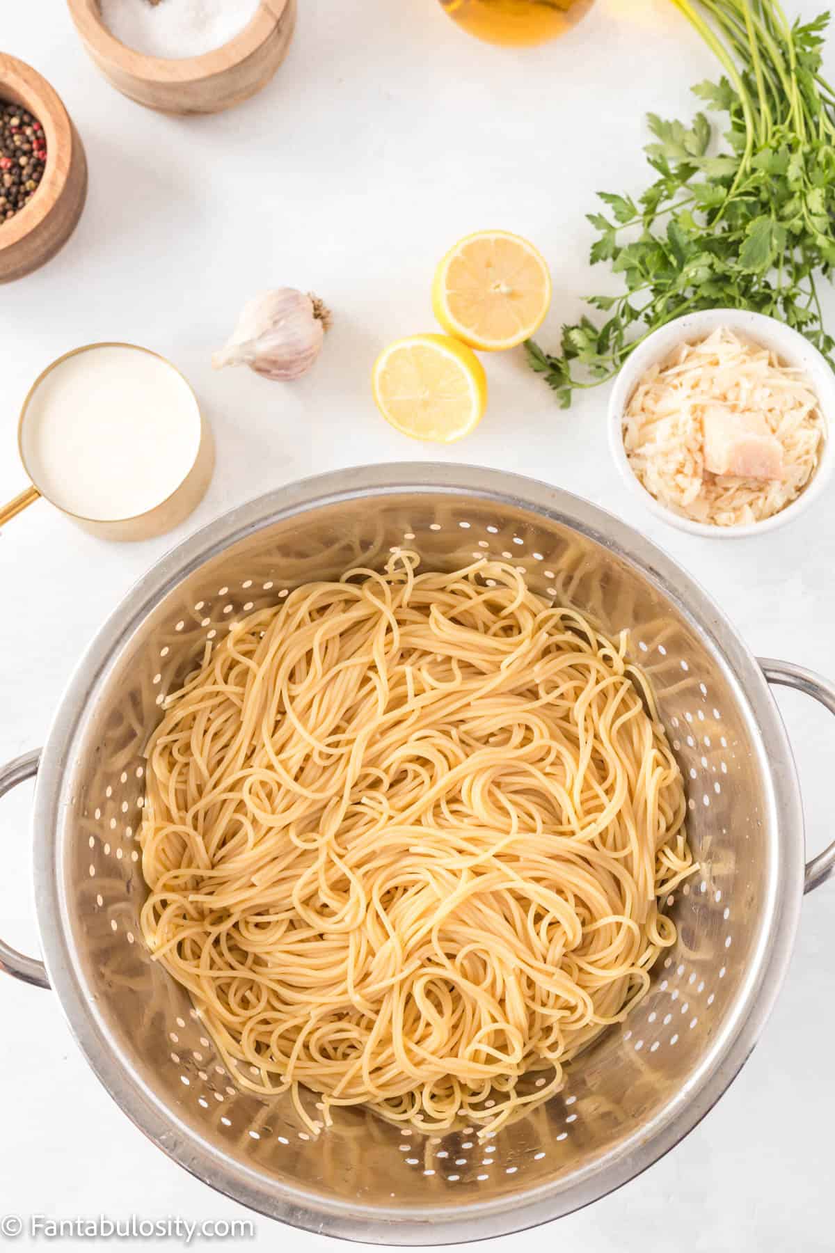 Cooked spaghetti noodles for Creamy Lemon Chicken Pasta are sitting in a colander surrounded by lemons, garlic, parsley and Parmesan cheese