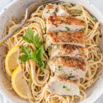 Close up photo of Creamy Lemon Chicken Pasta and a fork in a white speckled serving bowl garnished with lemon slices and fresh parsley