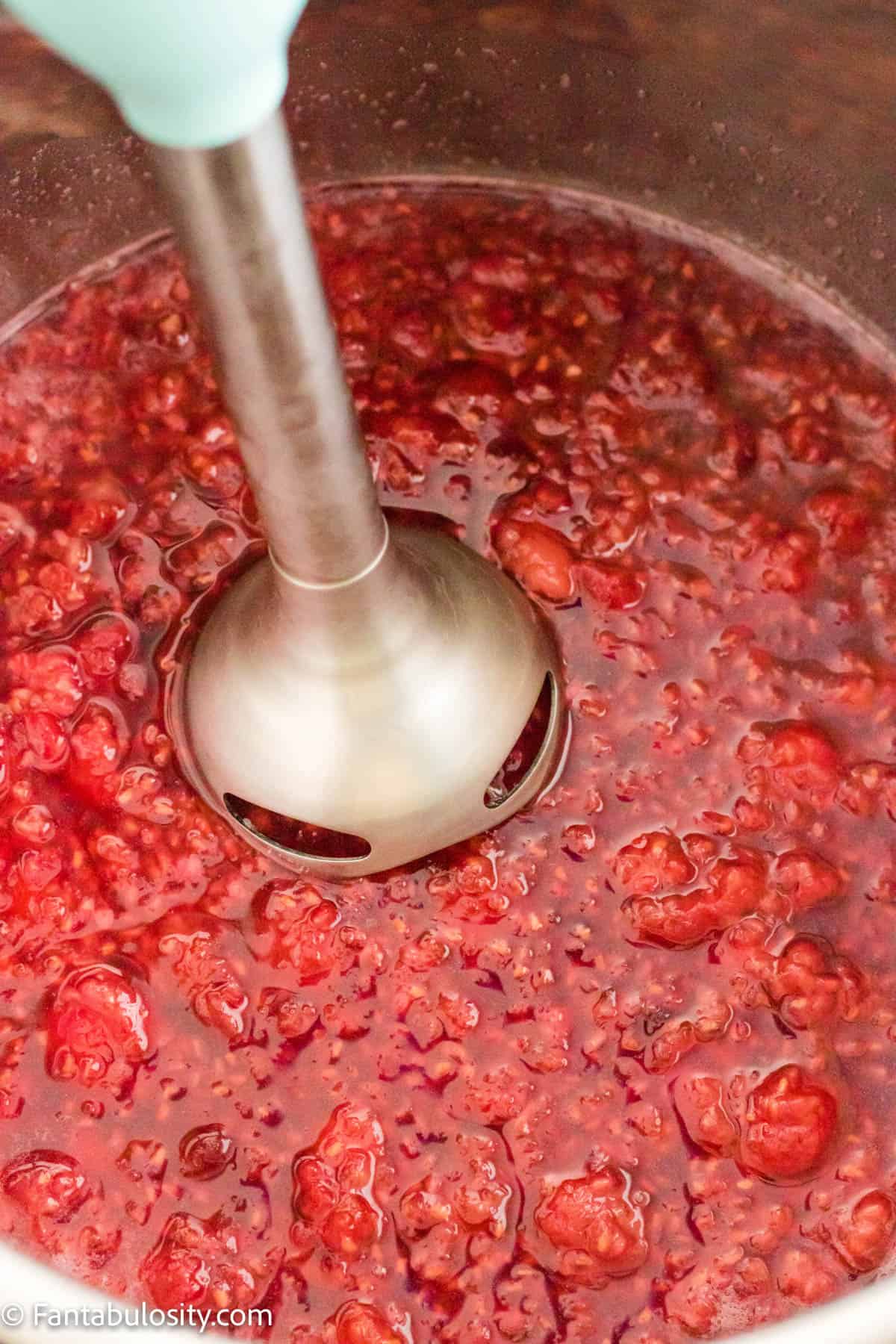 An immersion blender is hovering above a batch of Instant Pot raspberry jam ready to be blended