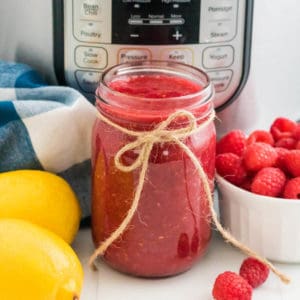 A mason jar of Instant Pot raspberry jam sits in front on of Instant Pot pressure cooker that is surrounded by lemons and berries