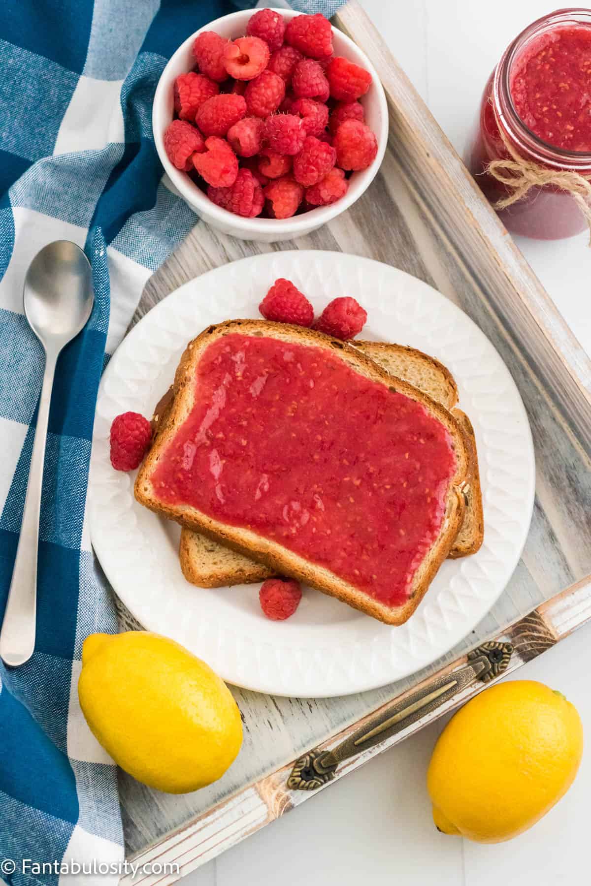 Vibrant red raspberry jam is spread on multigrain toast on a white plate. It is surround by lemons, a decorative gingham towel, fresh raspberries and a jar of jam