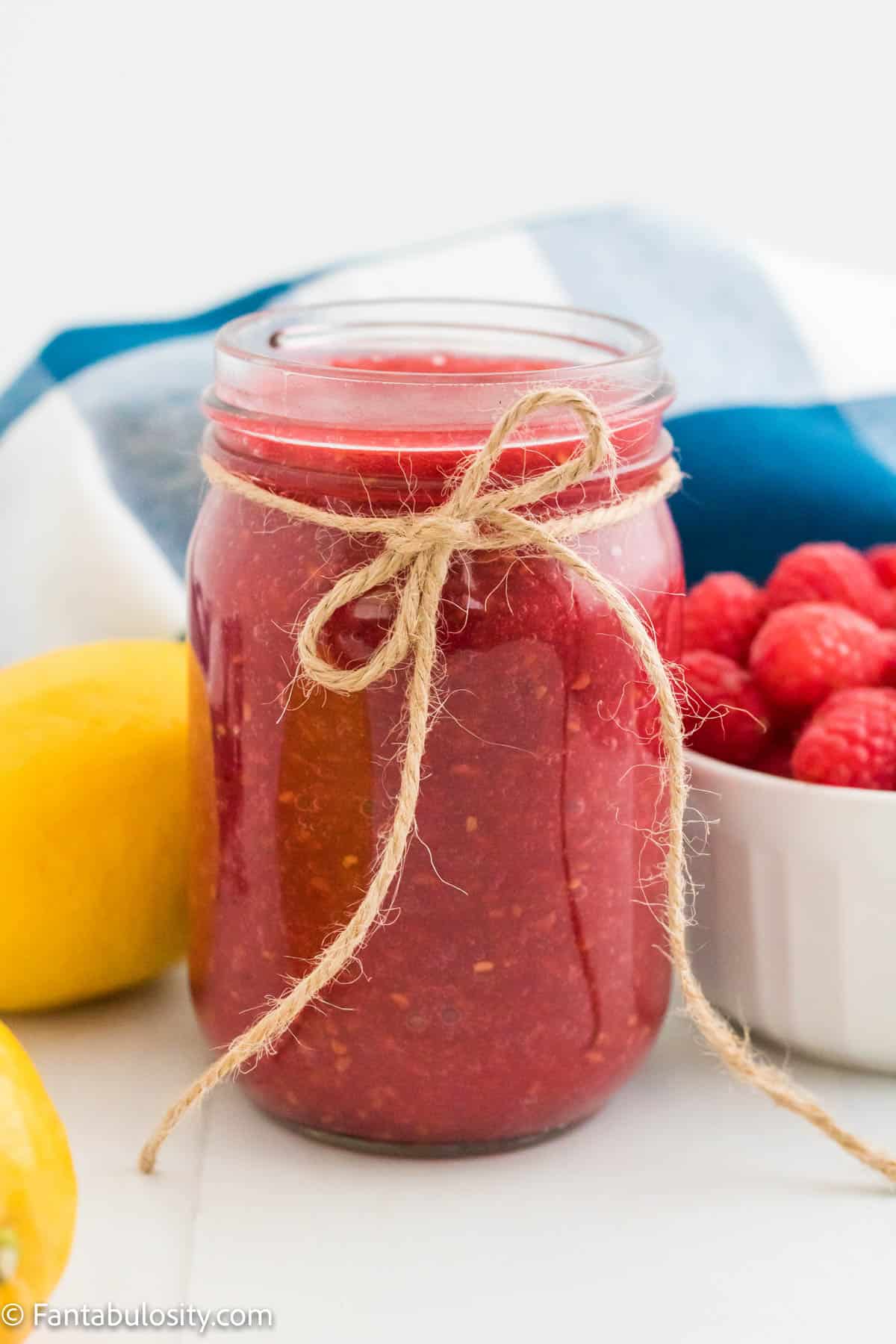A mason jar full of Instant Pot raspberry jam is centered and surrounded by lemons, a bowl of raspberries and a decorative gingham towel