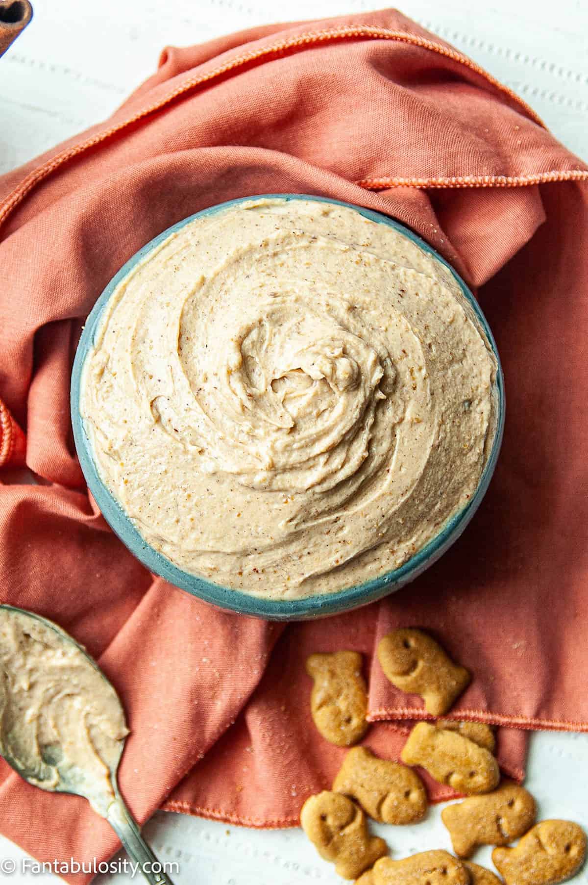 Creamy peanut butter yogurt dip is shown in the center of the photo on an orange background. There is a spoon on the left and graham crackers for dipping on the right.