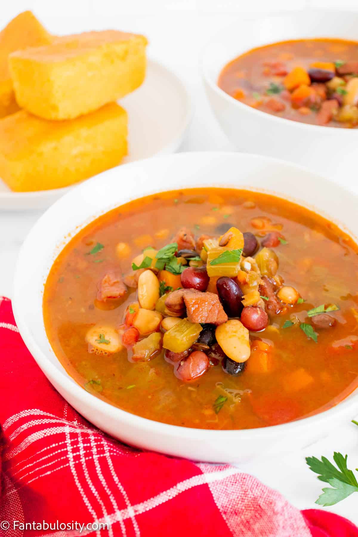 A bowl of 15 bean soup on a counter with a red napkin next to it and some cornbread on a plate behind it. 