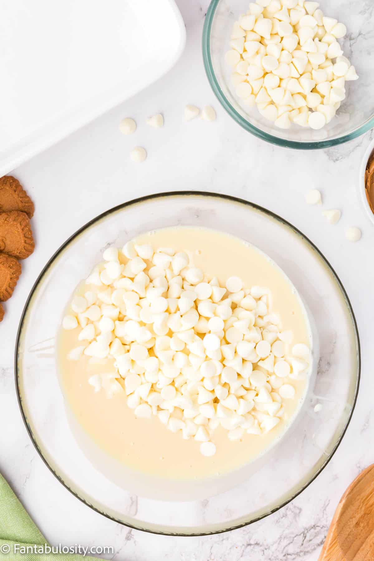 White chocolate chips have been added to a glass mixing bowl containing sweetened condensed milk and vanilla extract