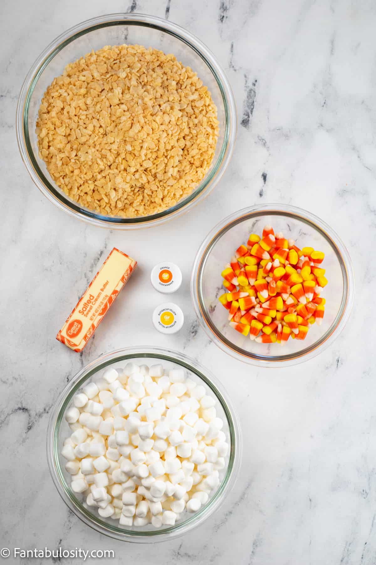 Bowls of the ingredients to make layered Candy Corn rice krispie treats, a stick of butter and food coloring are displayed on a white marbled background