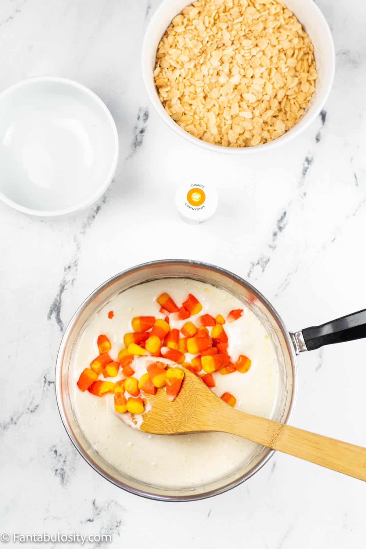 A sauce pan in centered and it contains a melted marshmallow mixture and candy corn. This is the beginning of the orange layer of candy corn rice krispie treats