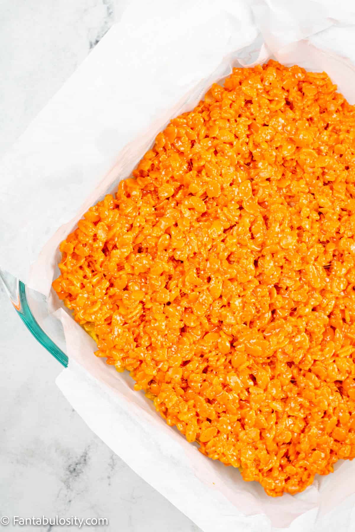 A layer of orange tinted rice krispie treat mixture has been pressed into a parchment paper lined 8x8 baking dish for candy corn rice krispie treats