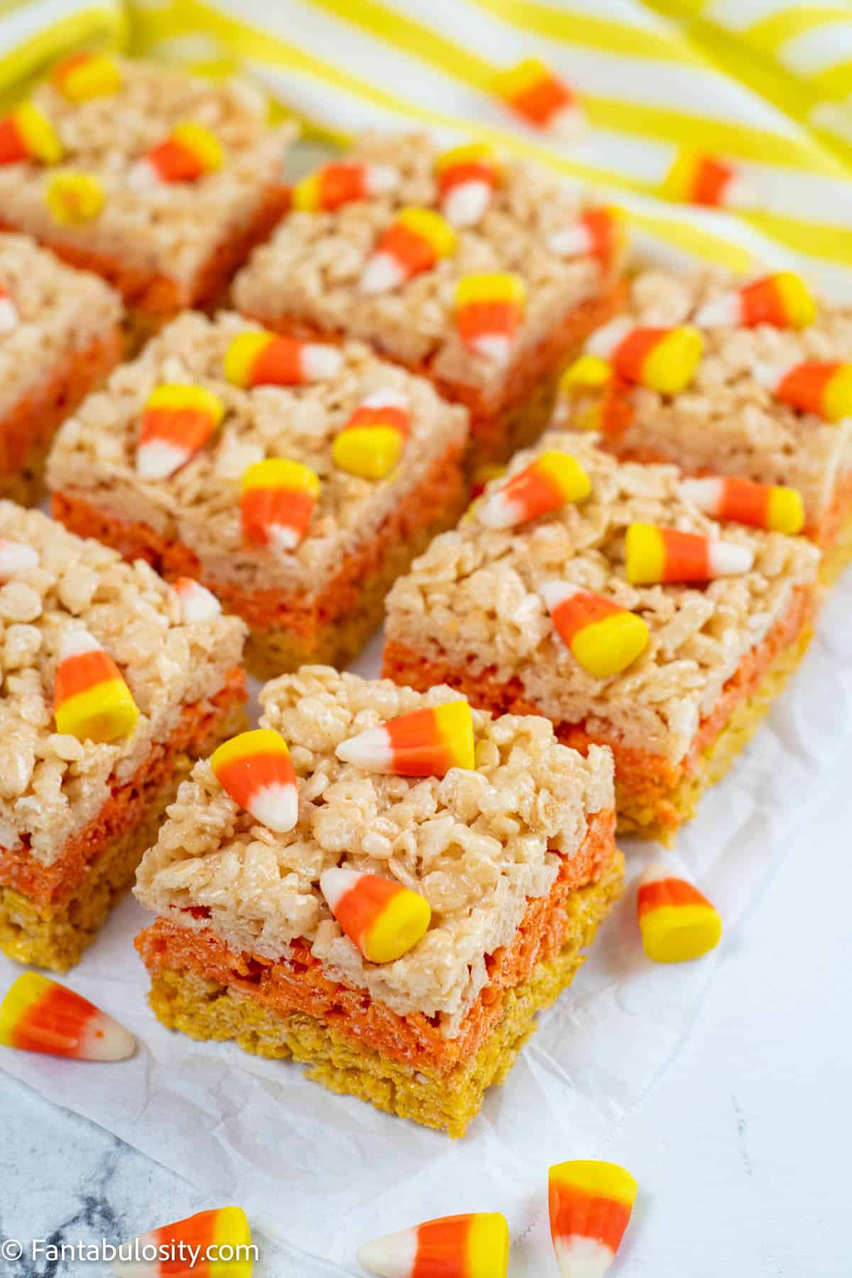 Squares of candy corn rice krispie treats are resting on parchment paper and surrounded by scattered candy corn
