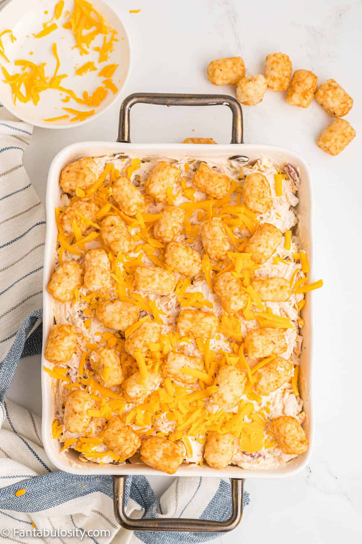 In a white baking dish, frozen tater tots top the unbaked cheesy chicken filling of a comforting casserole