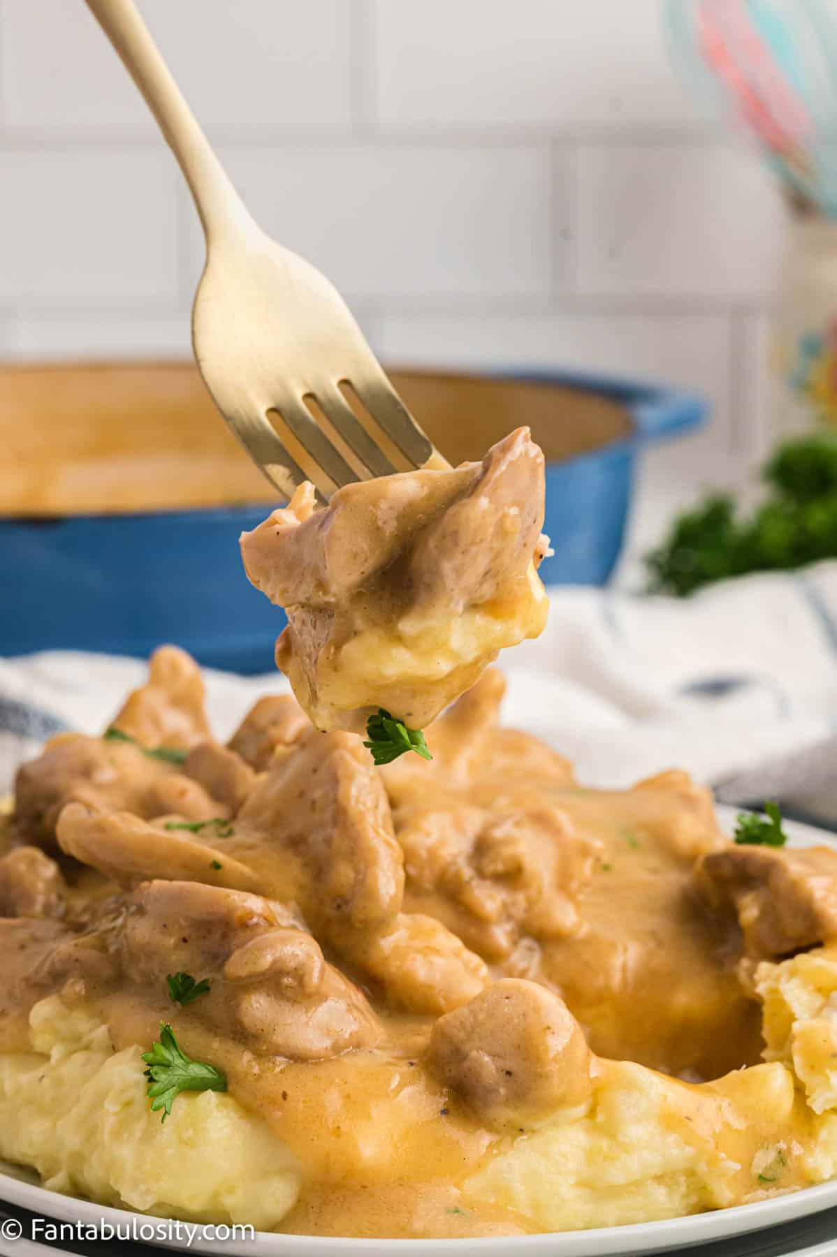 A gold fork has speared several pieces of tender chicken and is held over a plate of mashed potatoes smothered in the same creamy chicken