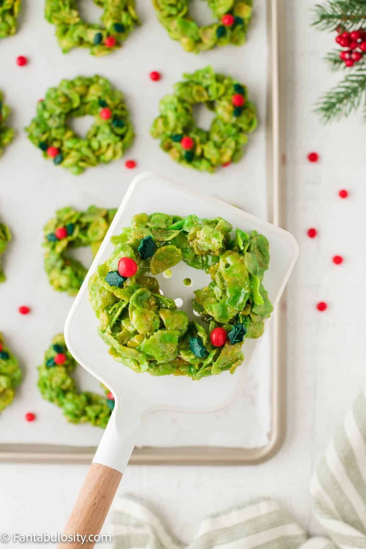 A white spatula holds a green cornflake wreath cookie above a baking sheet holding another batch of the festive holiday cookies