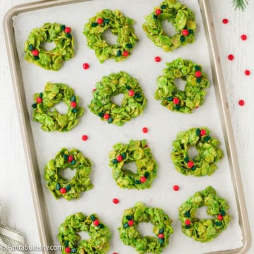 Bright green cornflake cookies dotted with bright red cinnamon candies fill a parchment lined baking sheet