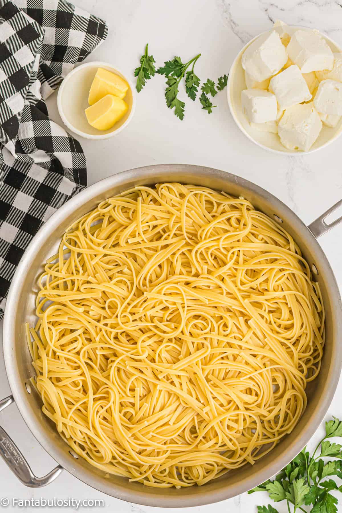 Boiled linguine noodles in a pot with butter, cream cheese, and parsley placed around the pot. 