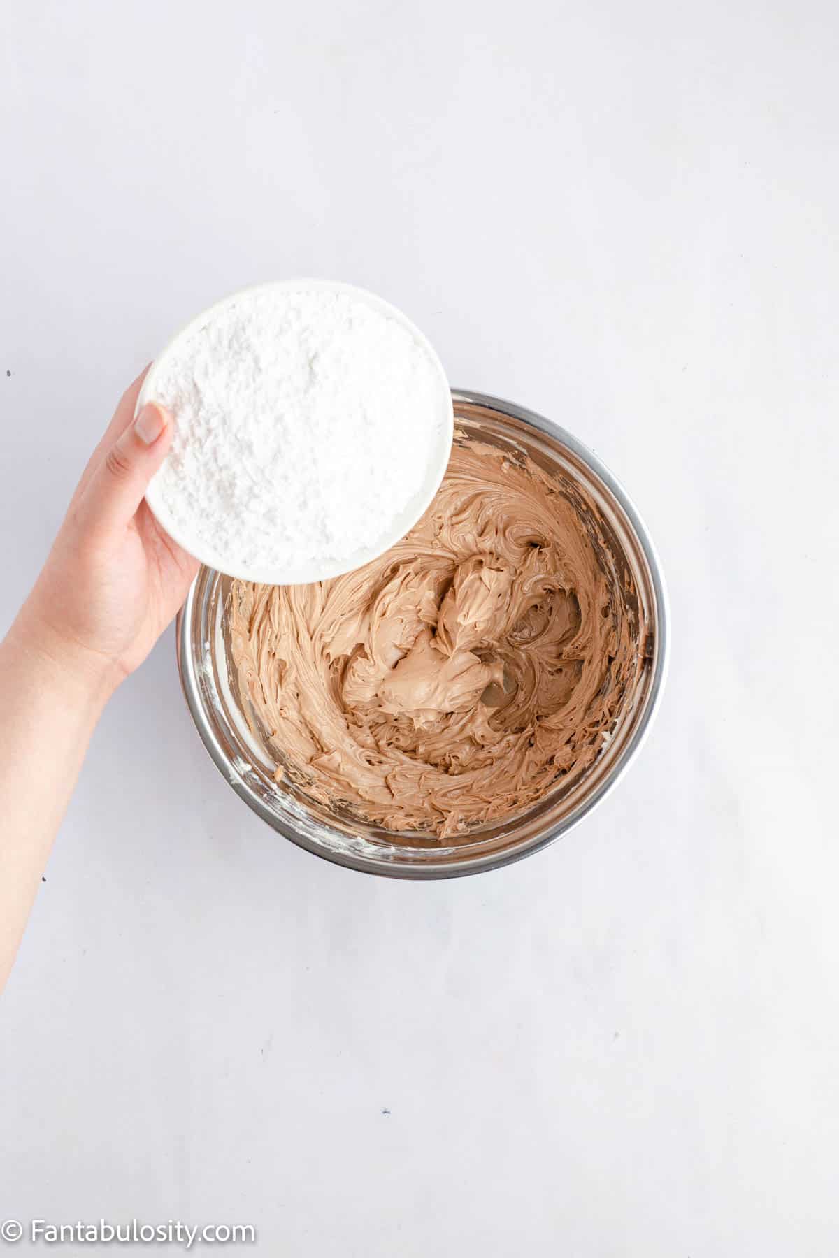 A hand is holding a bowl of powdered sugar ready to be added to a bowl of chocolate cream cheese frosting