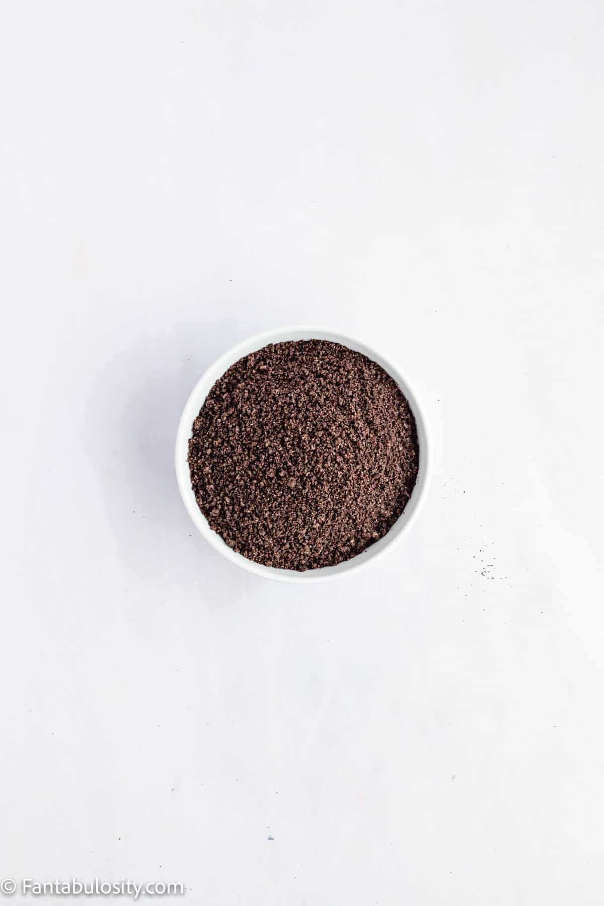 A bowl full of fine Oreo cookie crumbs is centered on a white background