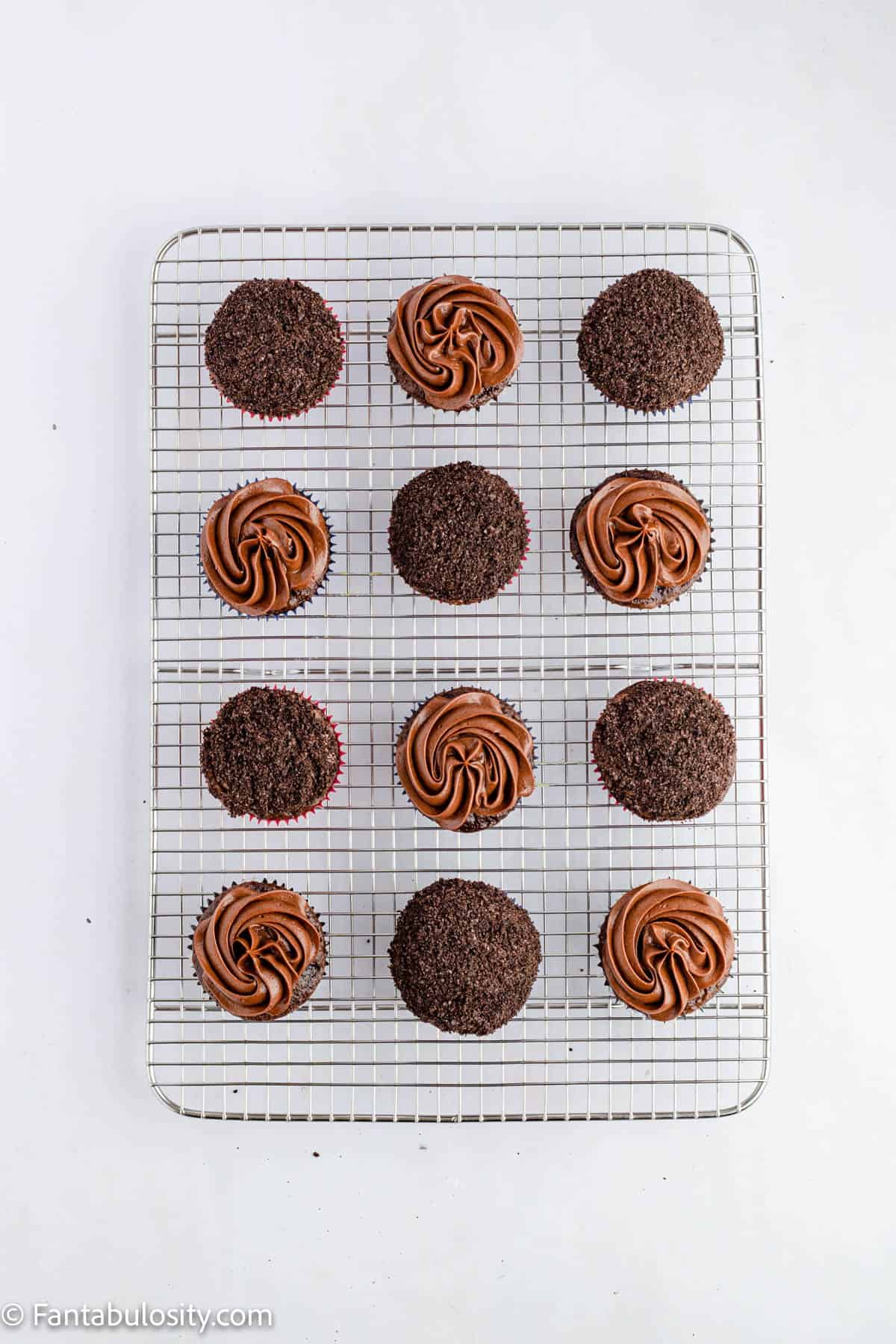 Chocolate cupcakes are displayed on a wire cooling rack. Half of them are frosting with chocolate frosting and half of them are topped with Oreo crumbs