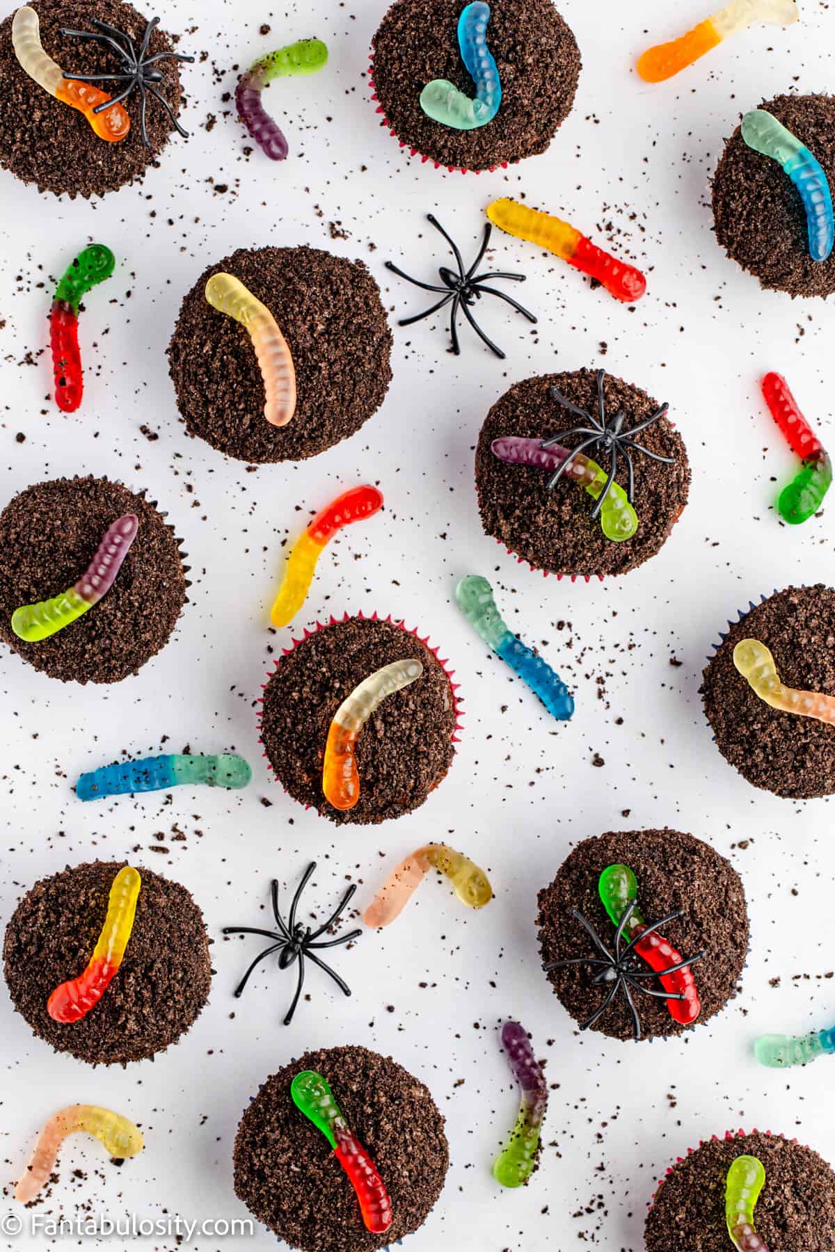 Frosted chocolate cupcakes are topped with Oreo crumbs and gummy worms, surrounded by plastic spiders, gummy worms and Oreo crumbs