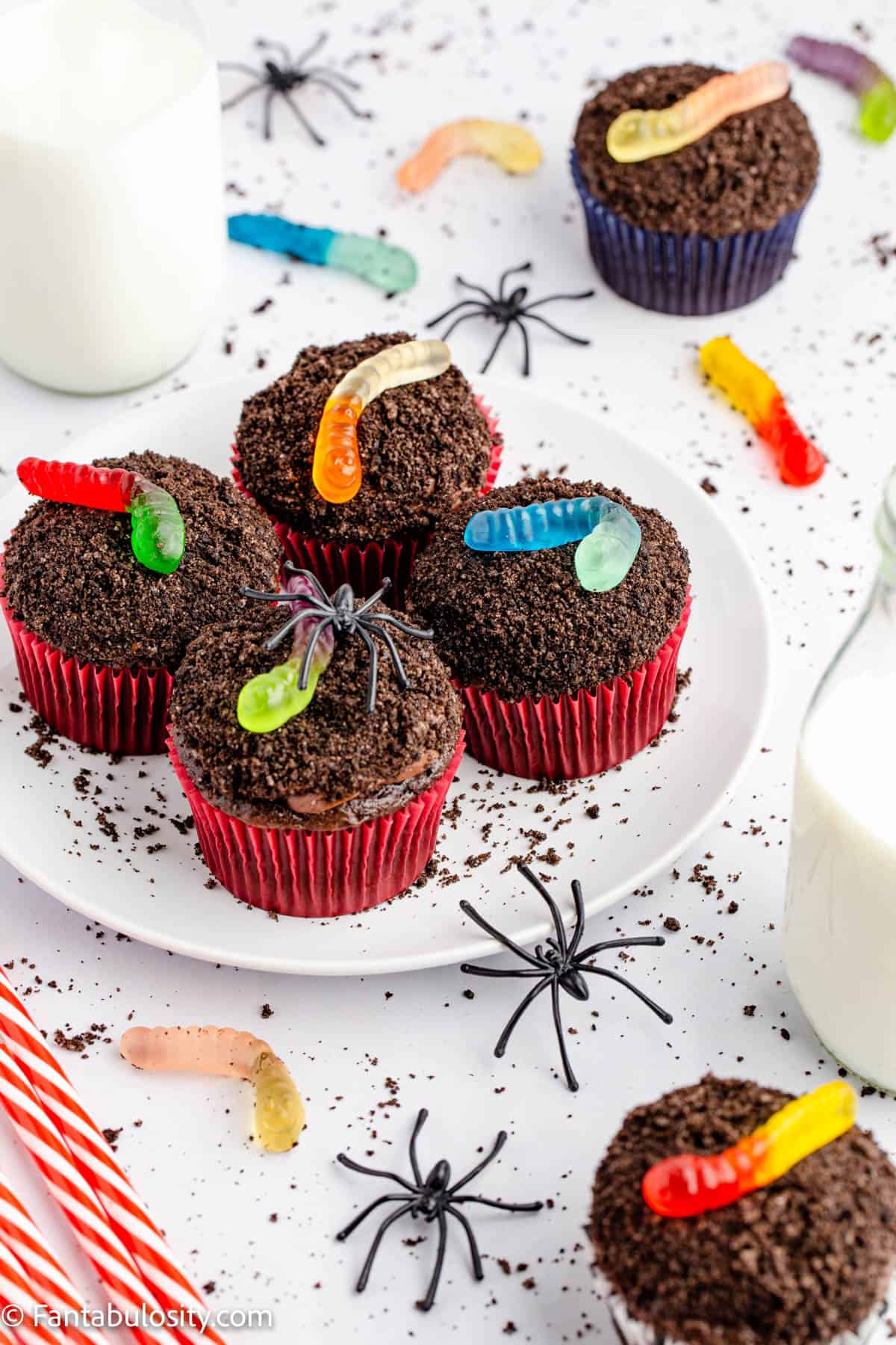 Frosted chocolate cupcakes are topped with Oreo crumbs and gummy worms, surrounded by plastic spiders, gummy worms and Oreo crumbs