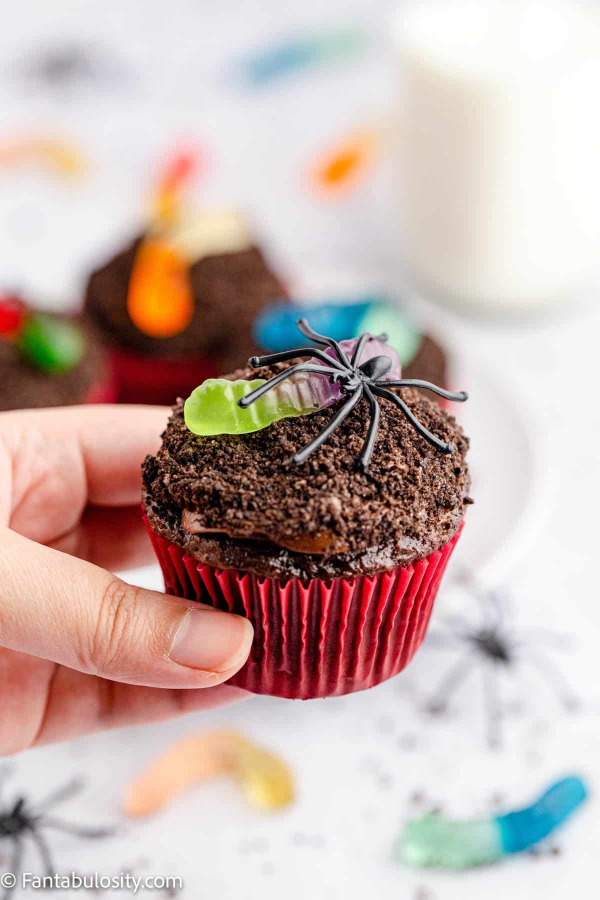 A hand is holding a chocolate frosted cupcake topped with Oreo crumbs and a colorful gummy worm
