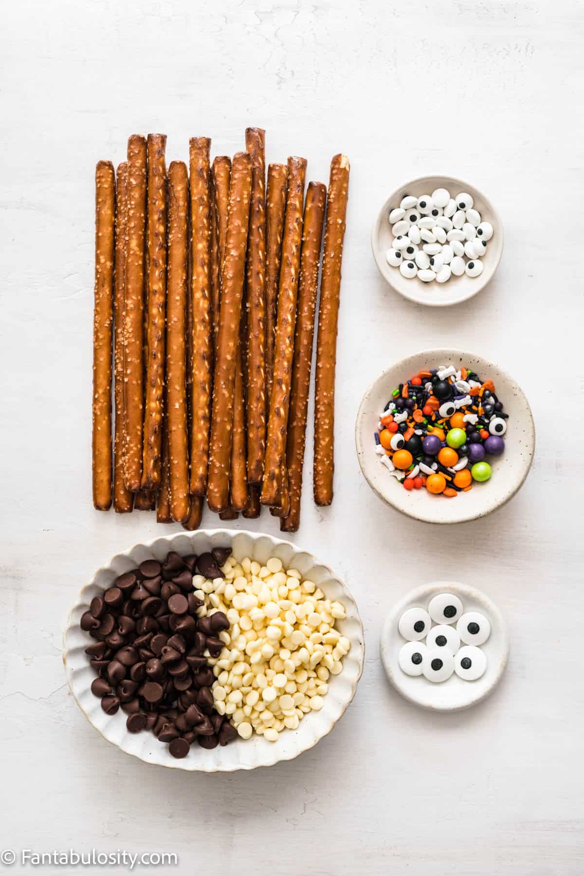 Pretzel rods, a bowl of chocolate chips, candy eyes and Halloween sprinkles are displayed on a white background