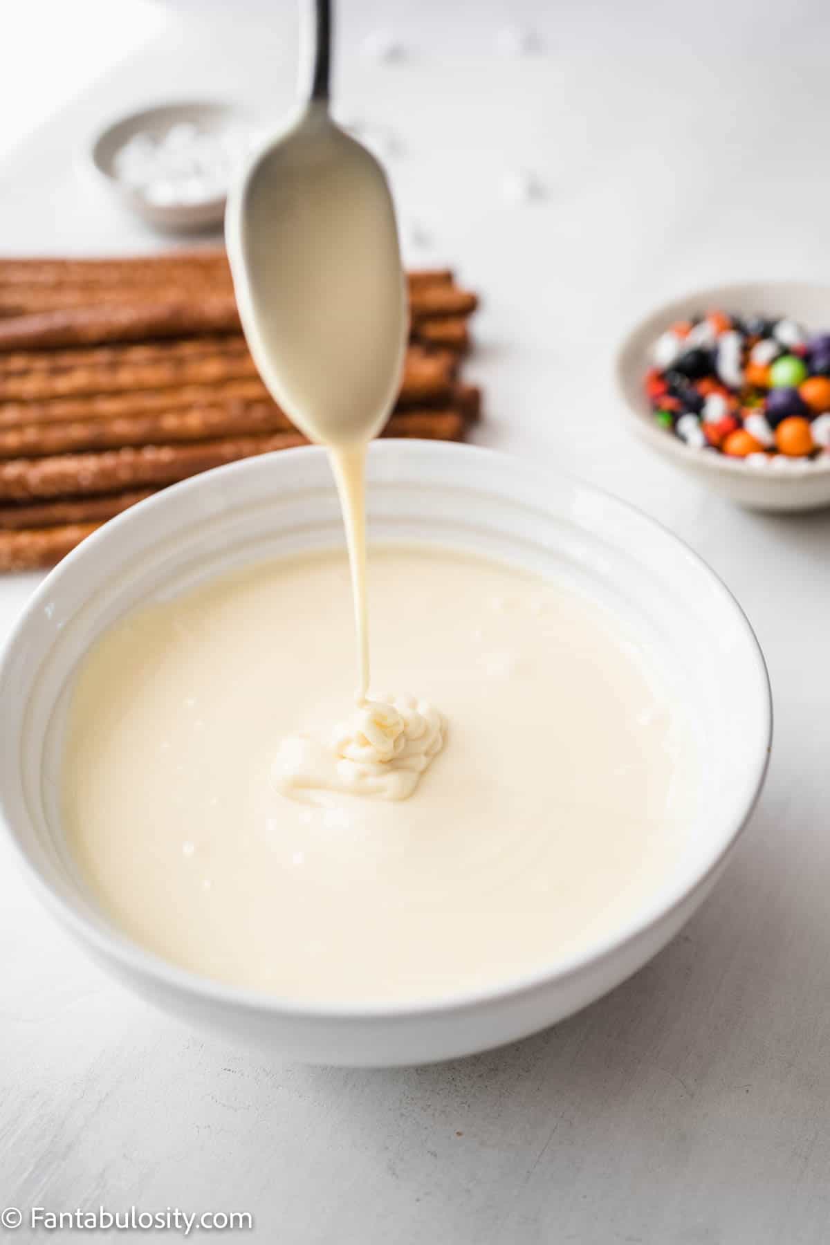 A bowl of melted white chocolate is in the center with a spoon being pulled from the chocolate. Halloween sprinkles and pretzel rods can be seen in the background.